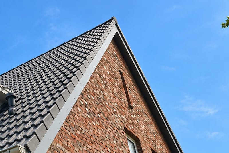 Steildach mit dunkler Ziegeldeckung, Ortgangblech und Dachentlüfter am Giebel eines Klinkerhauses vor blauem Himmel